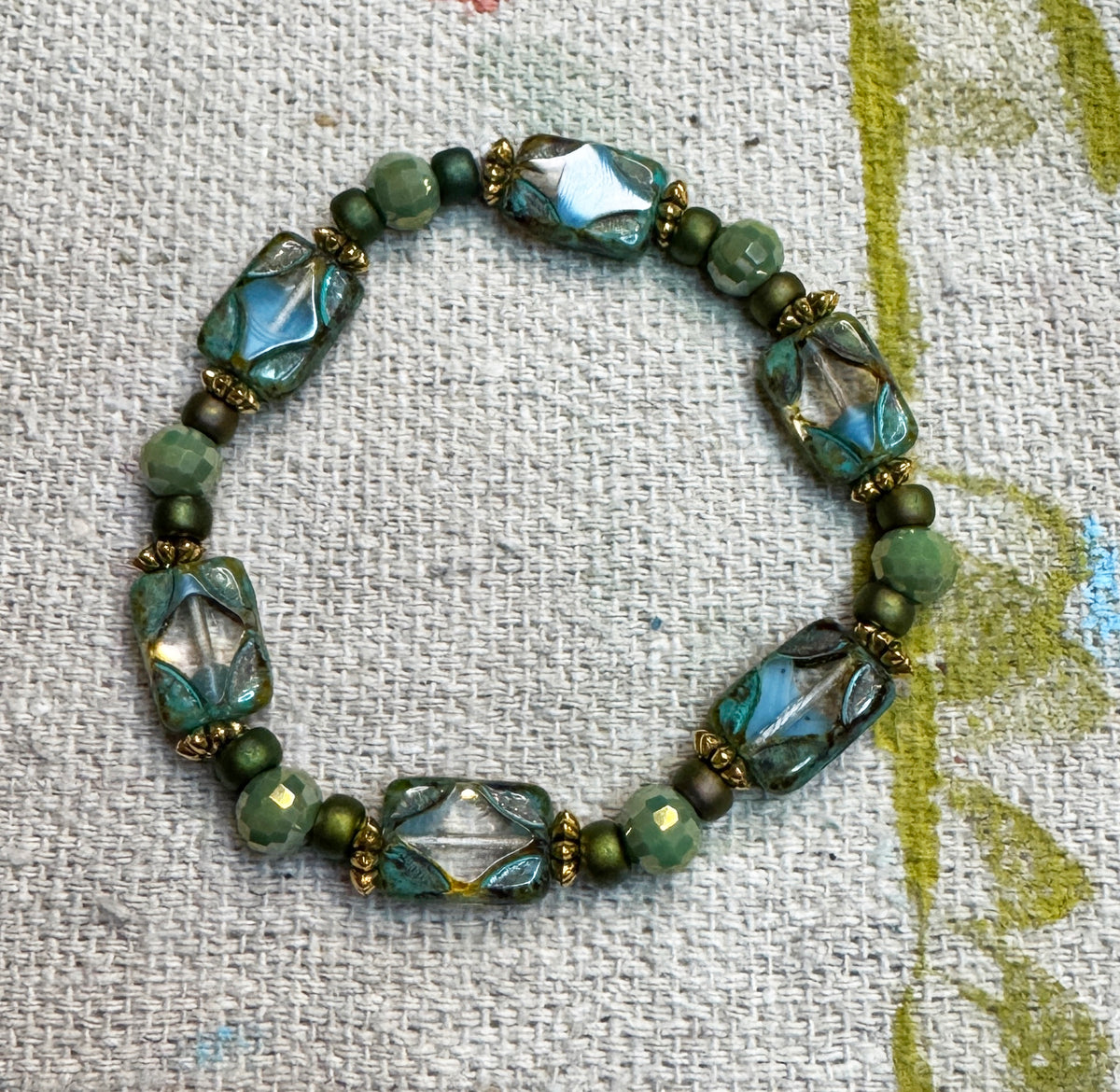 A handmade beaded stretch bracelet featuring green and blue-green Czech retangle table cut beads, amazonite facted beads, metallic green 6/0 seed beads and gold-toned accentss, displayed on a textured off-white linen fabric.