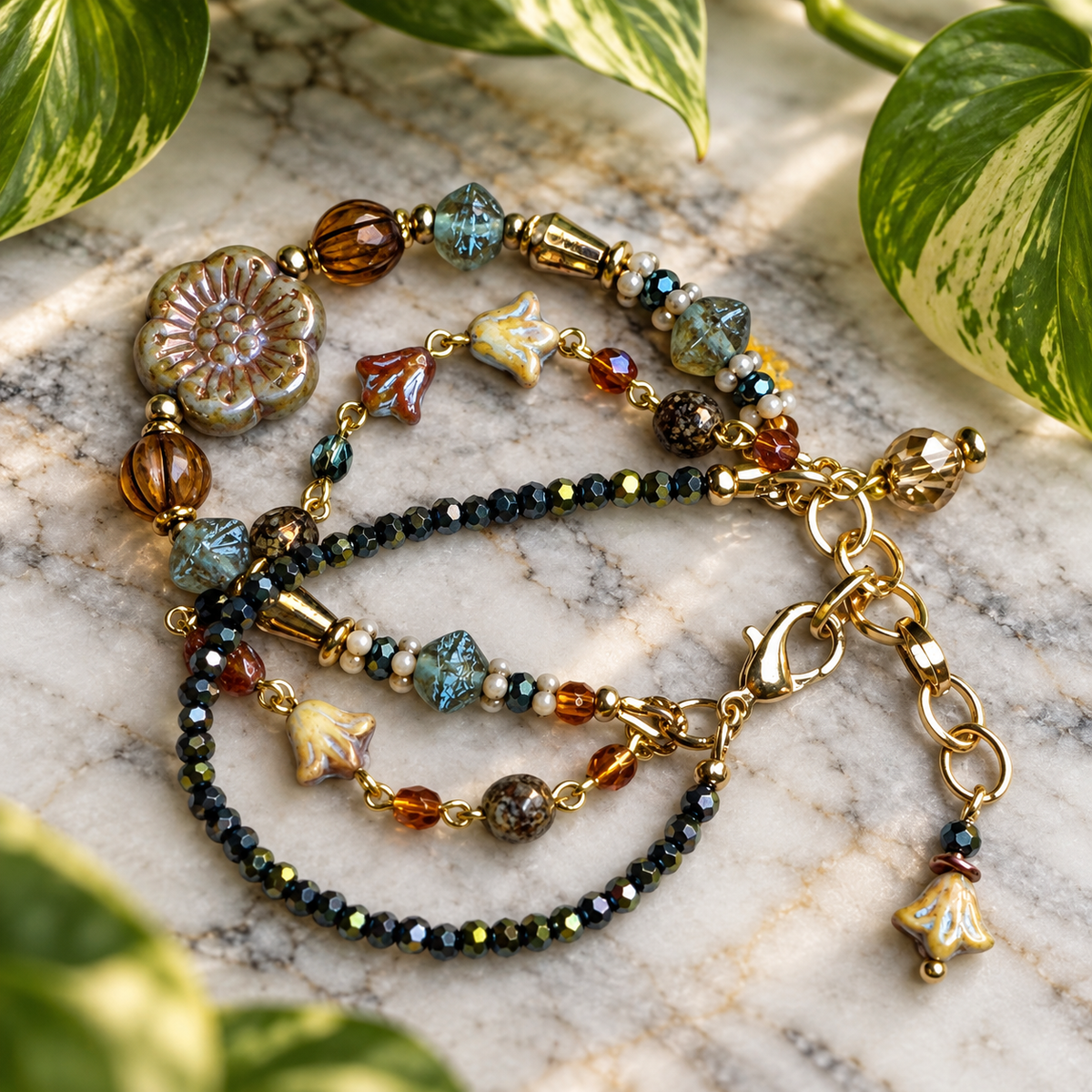 A 3 stranded beaded bracelet with gold-tone chain and clasp, featuring amber, blue, navy, and teal iridescent cube beads, displayed on a marble surface with green plant leaves.