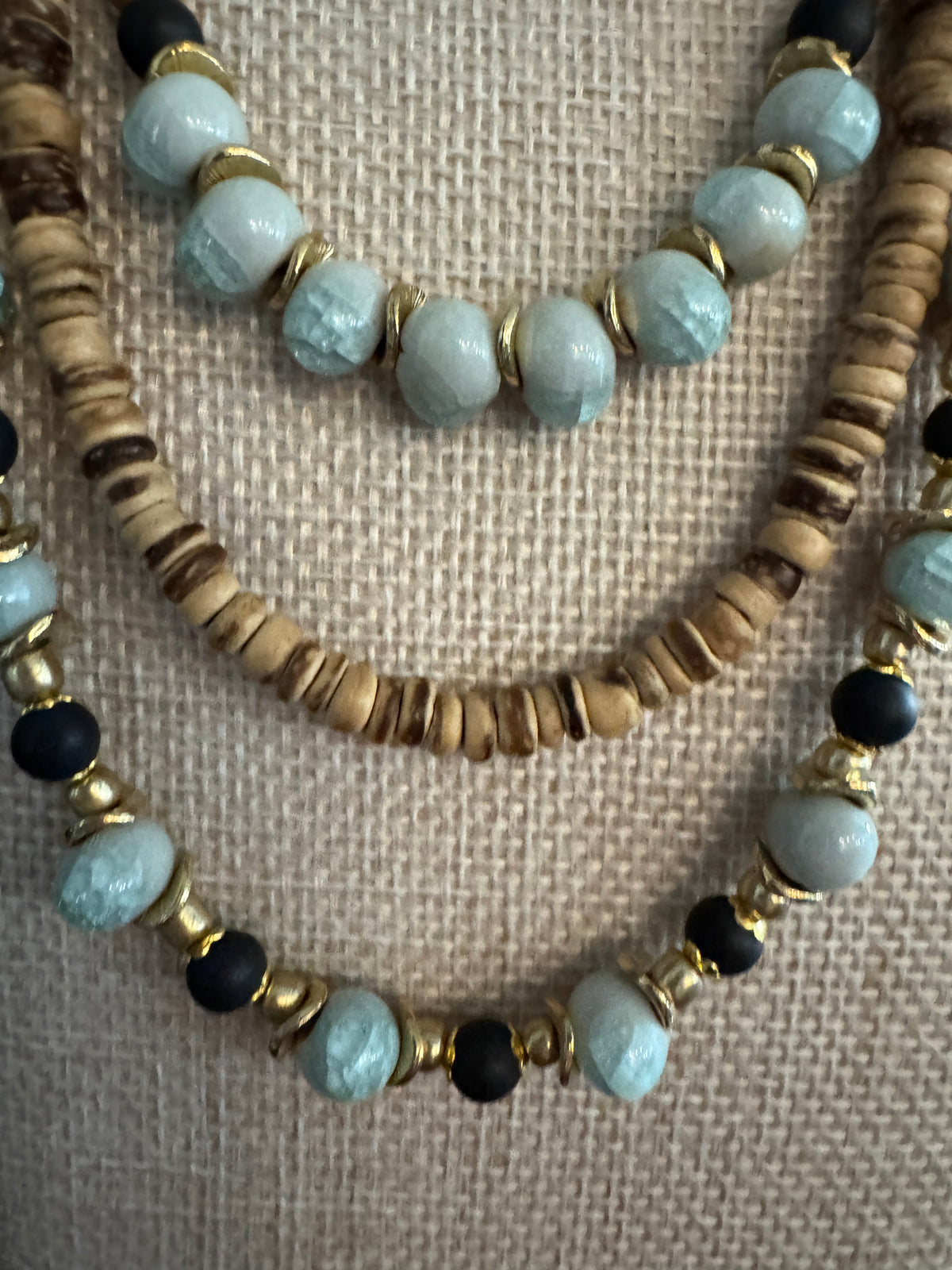 A close-up of a multi-strand beaded necklace displayed on a textured beige fabric background. The necklace features light blue marbled beads, dark brown wood-grain beads, small gold spacer beads, and dark charcoal black beads.