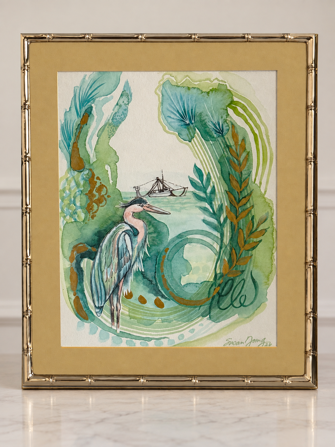 A framed watercolor painting featuring a a heron and aquatic plants, in the distance is a shrimp boat, set in a gold bamboo-style frame with a gold matching mat, displayed on a light marble surface.