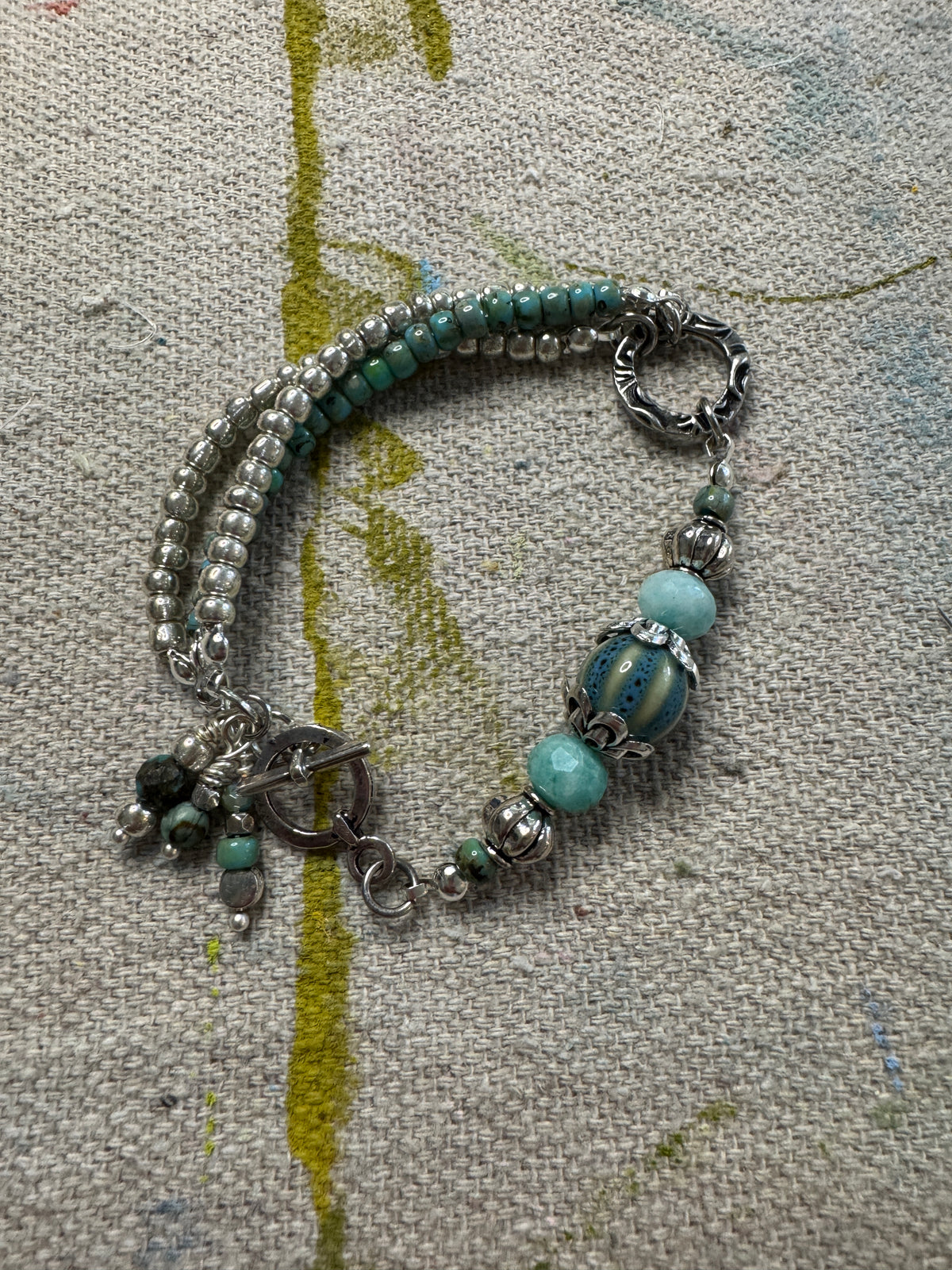 A handmade beaded bracelet with silver-toned metal beads, turquoise-colored round beads, and a toggle clasp, displayed on a textured beige fabric background.