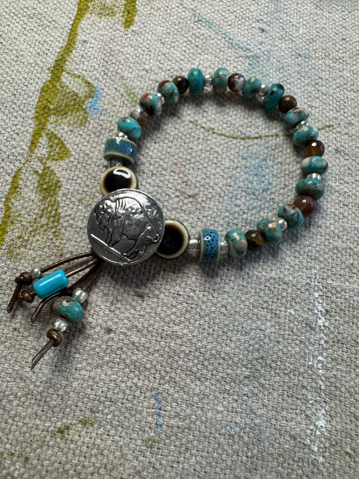 A beaded stretch bracelet featuring silver, brown, turquoise marbled beads, a central round silver-toned button charm, and dangling brown cord tassels with small turquoise and silver beads, displayed on a textured beige linen fabric.