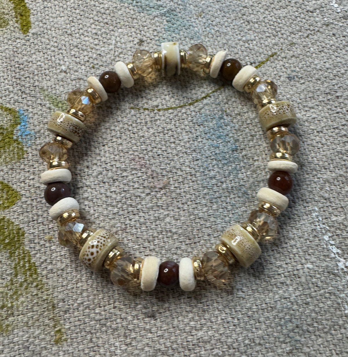A beaded stretch bracelet featuring cream-colored ceramic beads, wooden beads, faceted clear crystal beads, and dark brown beads, displayed on a textured gray fabric background.