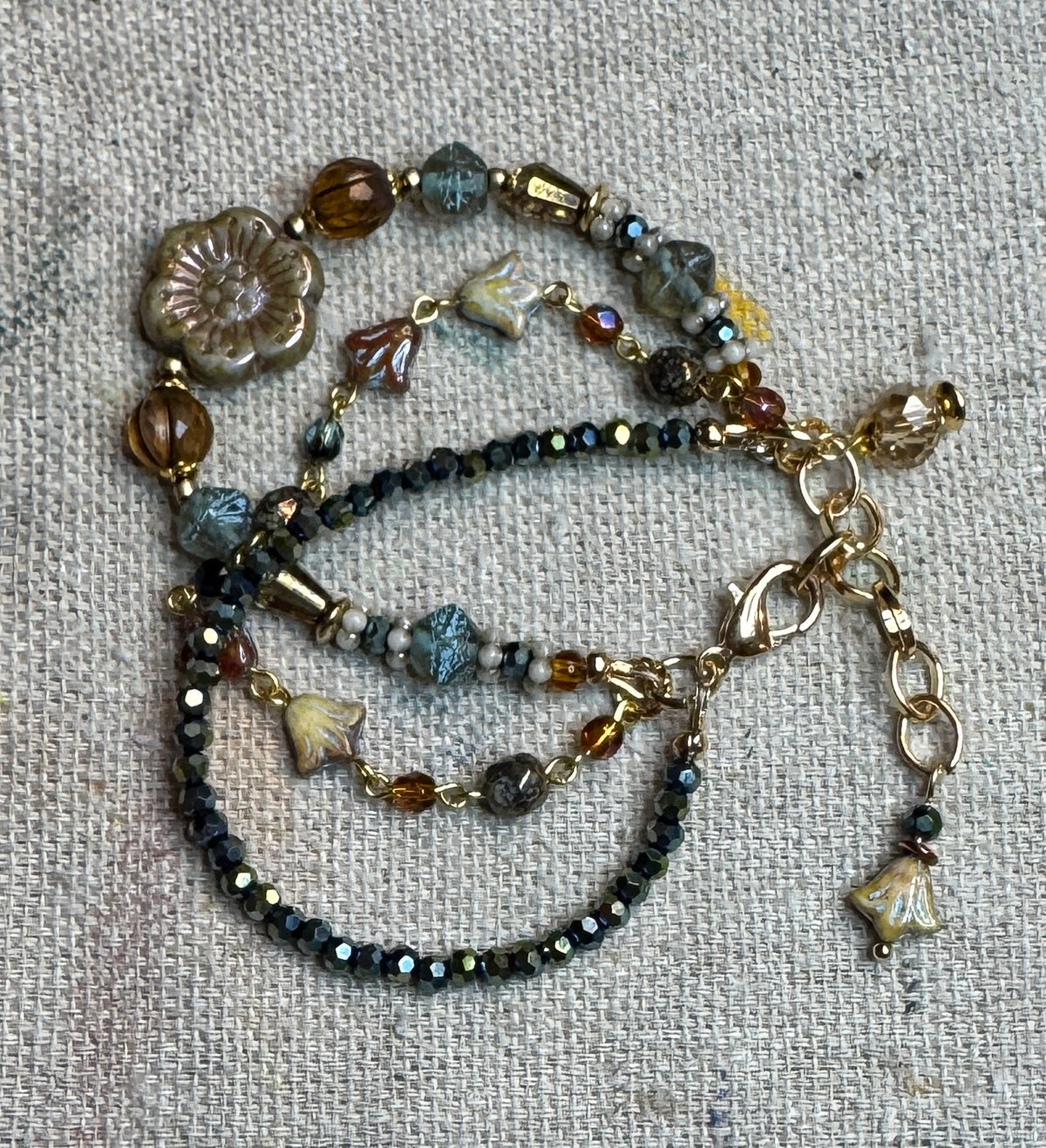 A 3 stranded beaded bracelet with gold-tone chain and clasp, featuring amber, blue, navy, and teal iridescent cube beads, laid flat on a textured linen background.