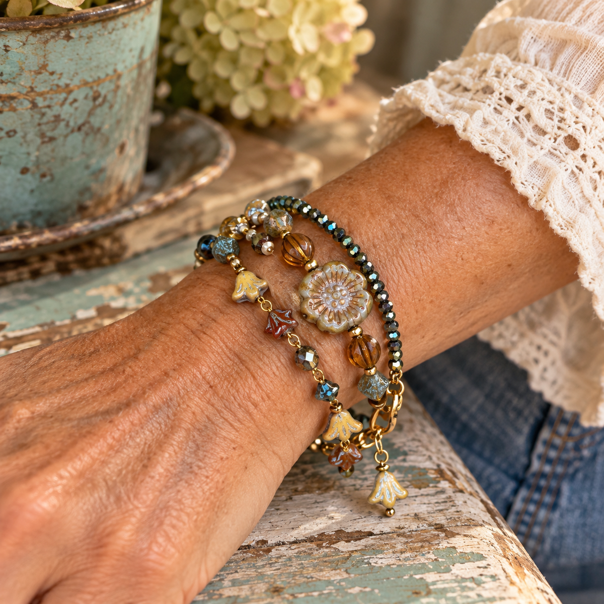 A close-up of a person's wrist wearing three layered bohemian-style beaded and charm bracelets, resting on a rustic distressed wooden table with a potted plant in the background.