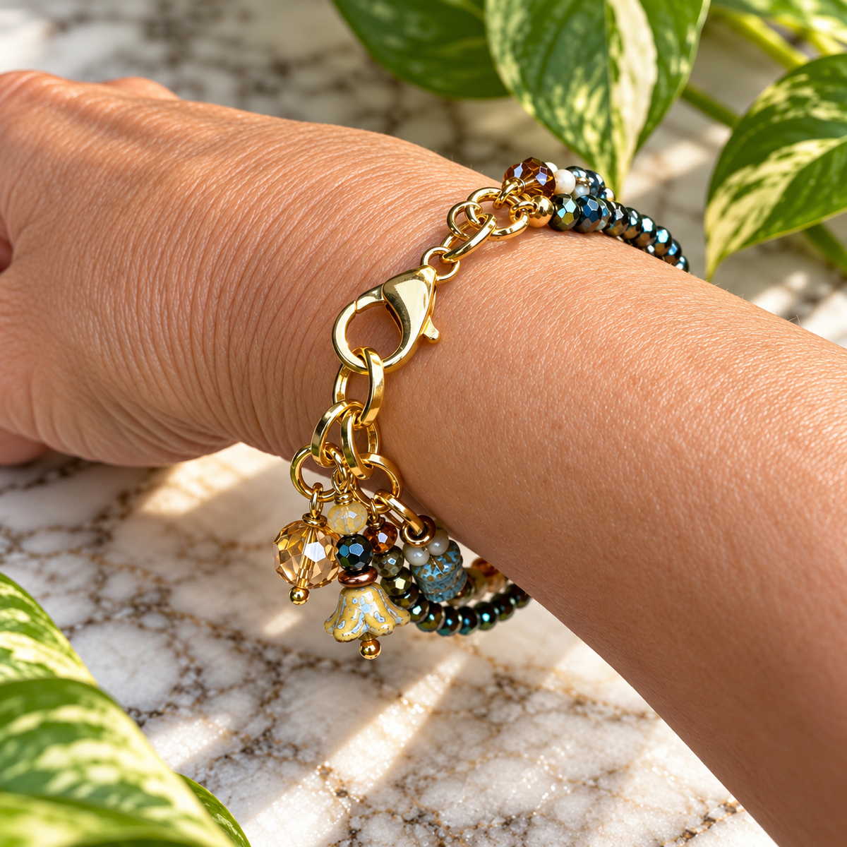 A gold-tone charm bracelet with colorful beads and charms, shown worn on a person's wrist against a marble surface with green leaves.