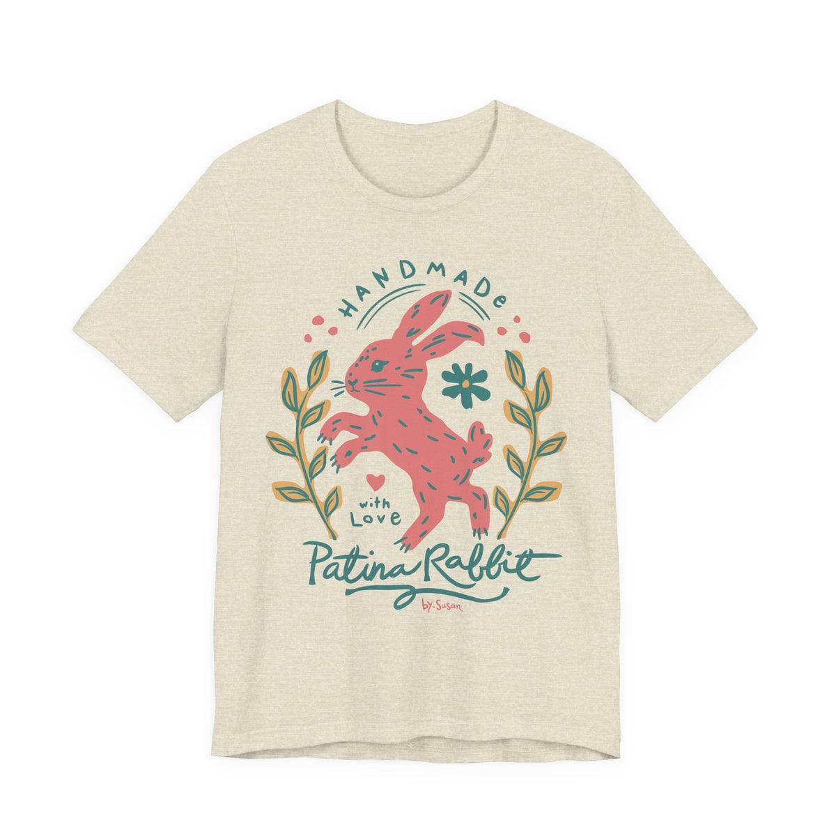 A light heather beige t-shirt featuring a colorful graphic of a pink rabbit with the text 'HANDMADE with Love Patina Rabbit' and decorative leaves.