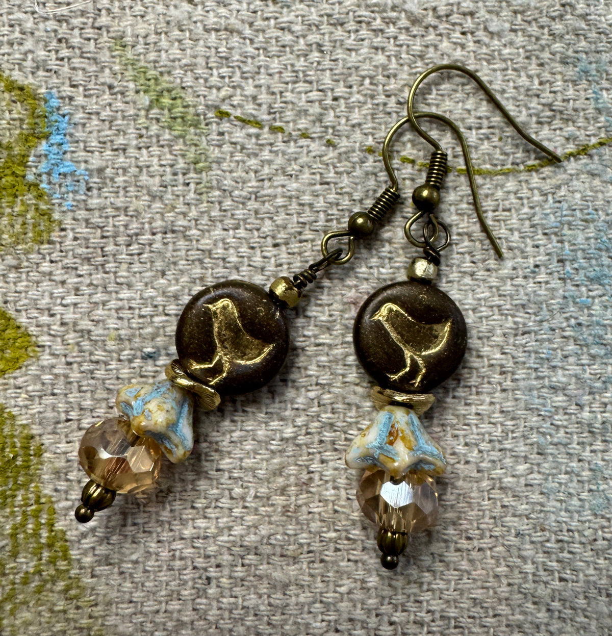 A pair of handmade dangle earrings with dark brown round beads featuring a gold bird design, hanging from bronze-toned wires, and accented with faceted light blue and clear beads, displayed on a textured beige fabric background.