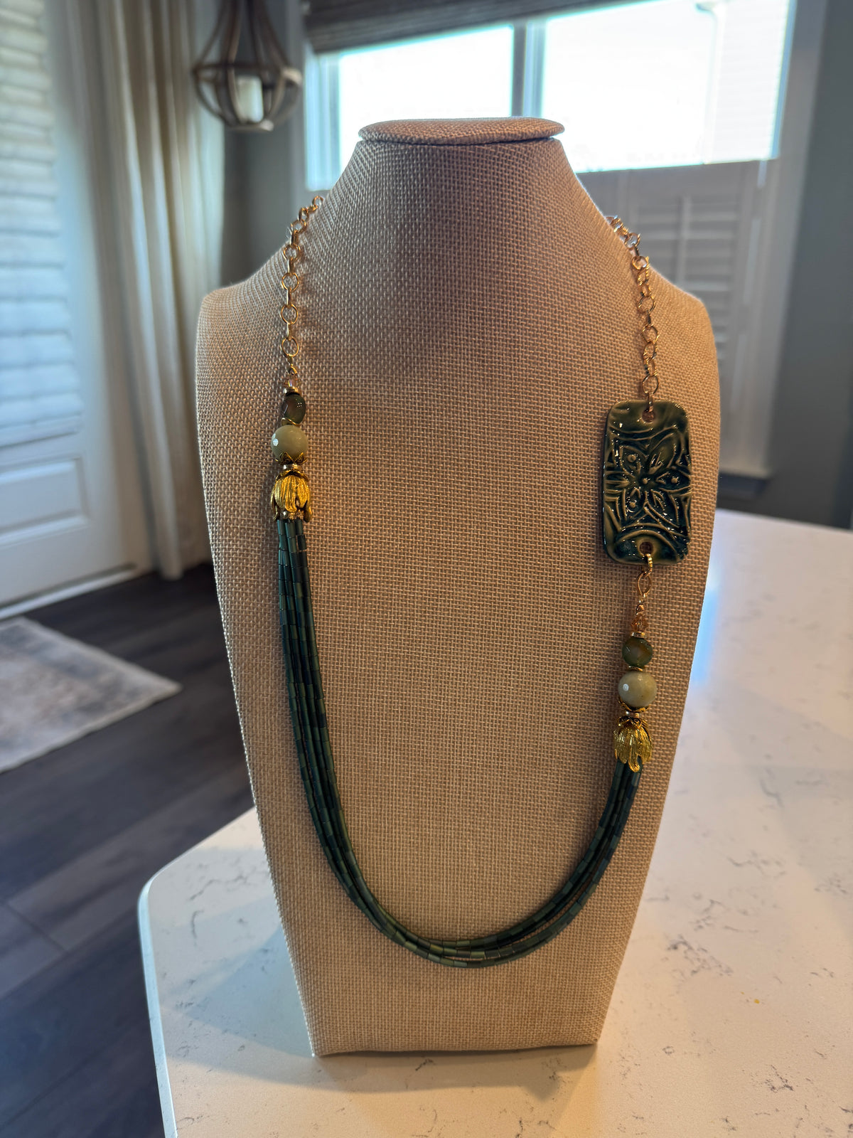 A long statement necklace displayed on a beige fabric-covered jewelry bust. The necklace features a gold-tone chain, a large rectangular dark green carved pendant, green beads, and a long strand of dark green cord.