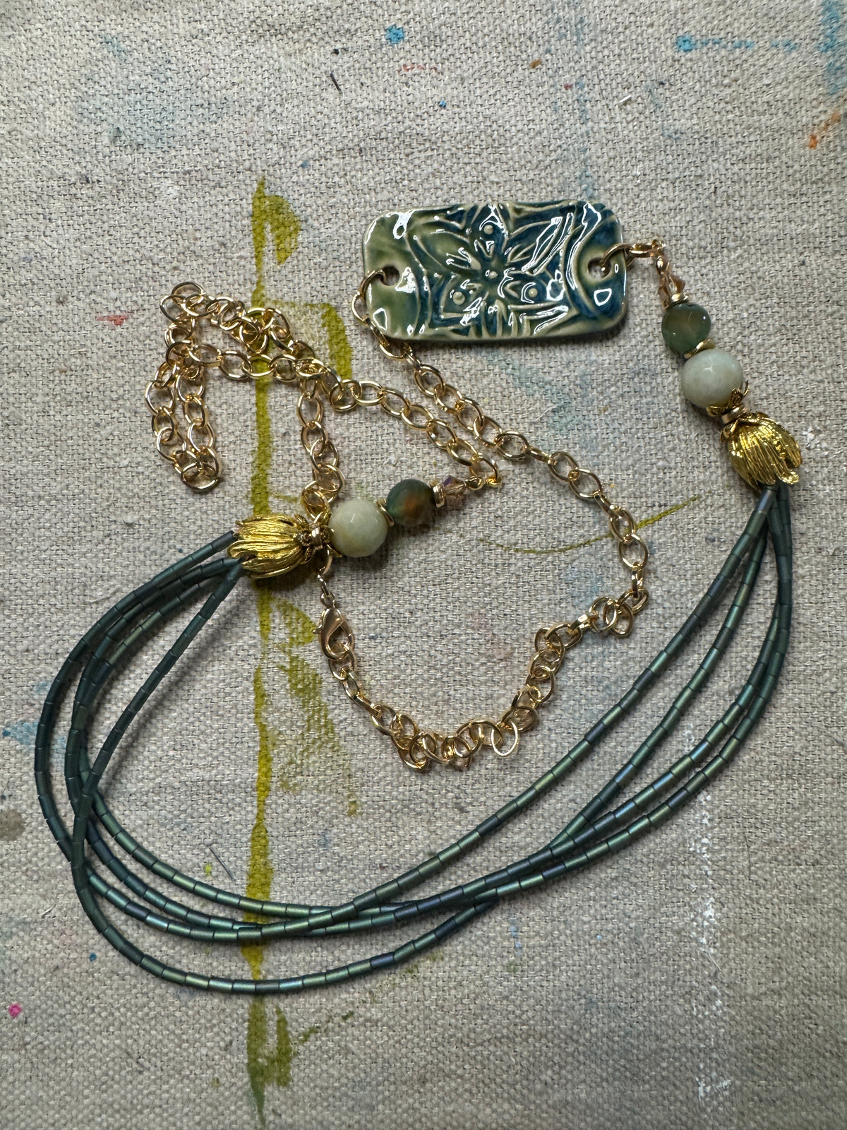 A handmade multi-strand necklace featuring dark teal-green cords, a gold-tone chain, a carved rectangular pendant with a butterfly motif, and decorative beads, laid out on a textured beige canvas background.