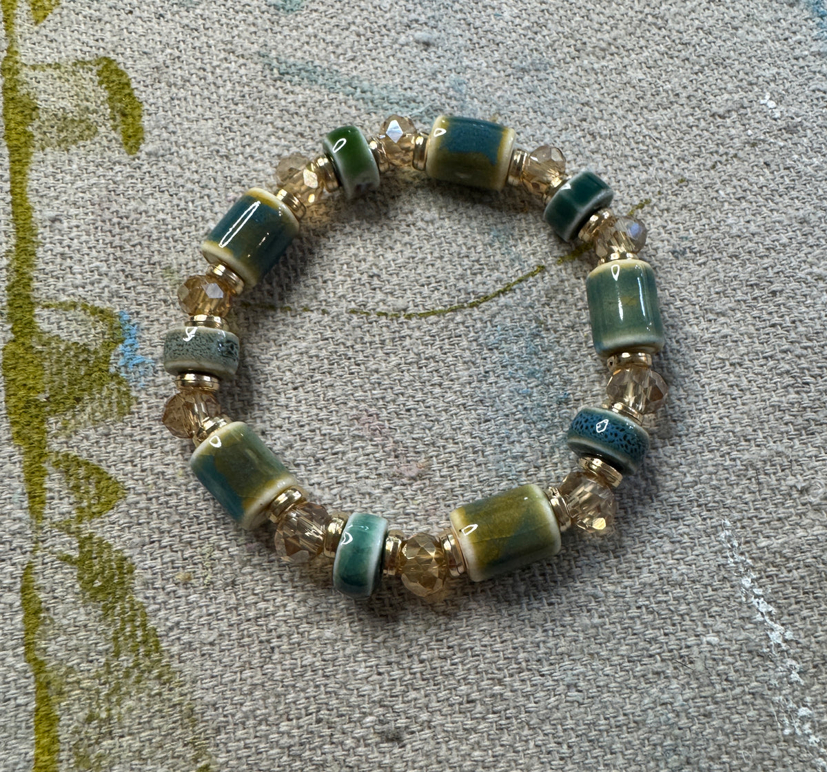 Seagrass & Moss stretch bracelet with moss green and deep teal ceramic beads, champagne glass beads, and gold accents spacer beads laying on an artisan drop cloth.