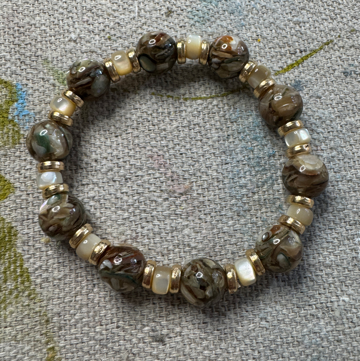 A beaded stretch bracelet featuring marbled brown lampwork beads, cream-colored spacer beads, and gold metal spacers, displayed on a textured gray fabric background.