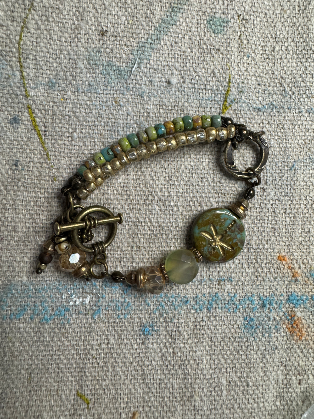 A handmade beaded bracelet with a toggle clasp, featuring a mix of green, blue, and gold-toned beads, a large round green stone, and a dragonfly bead motif on a textured linen background.