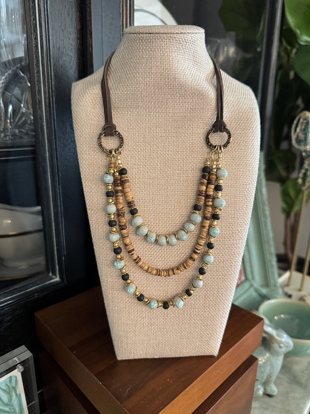A multi-strand beaded necklace displayed on a beige fabric jewelry bust. The necklace features light blue-green beads, natural wood beads, dark brown beads, and dark metal circular ring connectors.