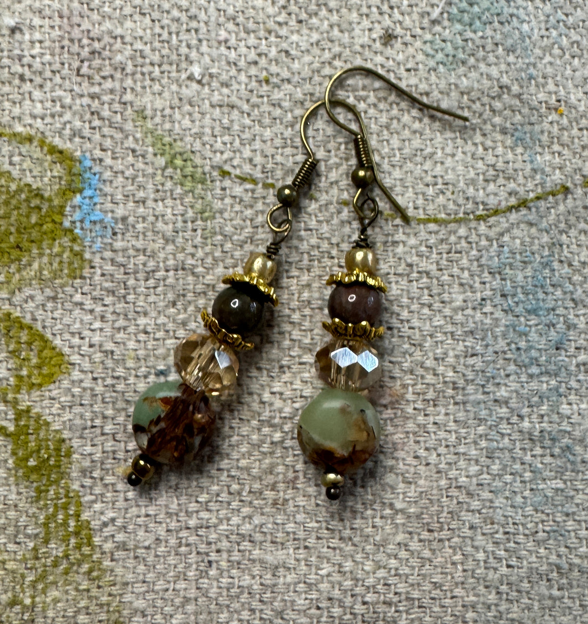 A pair of handmade dangle earrings with bronze-toned metal hooks and beads, featuring a faceted clear crystal, a dark brown bead, a round light green bead, and a marbled brown bead, displayed on a textured beige fabric background.