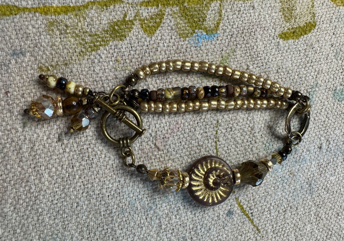 An antique-style beaded bracelet with bronze-toned metal components, including a sunburst shell charm, a toggle clasp, and multiple strands of beads in gold, brown, and black, displayed on a textured beige fabric background.