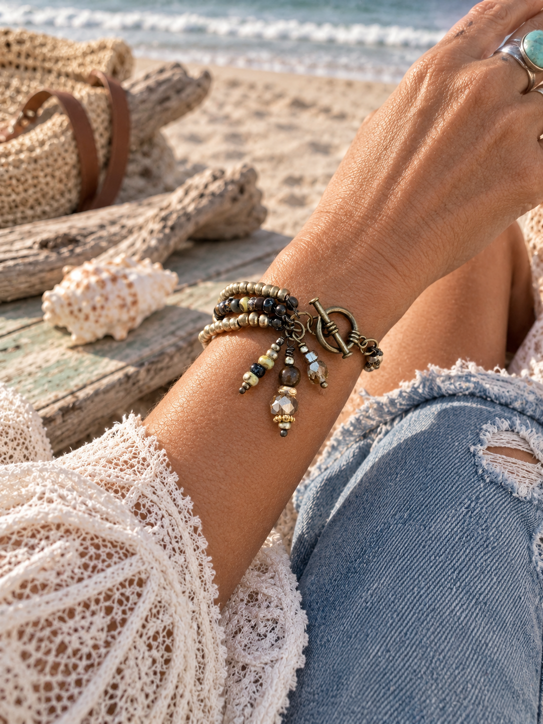 A close-up lifestyle shot of a person's wrist wearing a bohemian beaded bracelet with dangling charms, set against a beach background with a straw bag, seashell, and ocean.