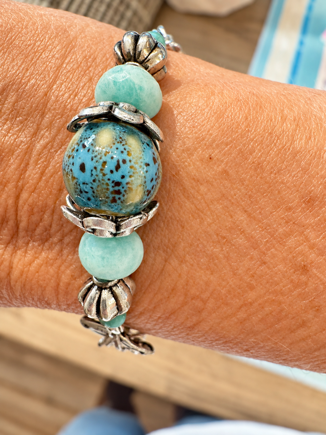 A close-up of a beaded bracelet worn on a person's wrist. The bracelet features a large central turquoise-blue beaded sphere with brown speckles, flanked by light aqua round beads and silver-toned decorative metal spacers.