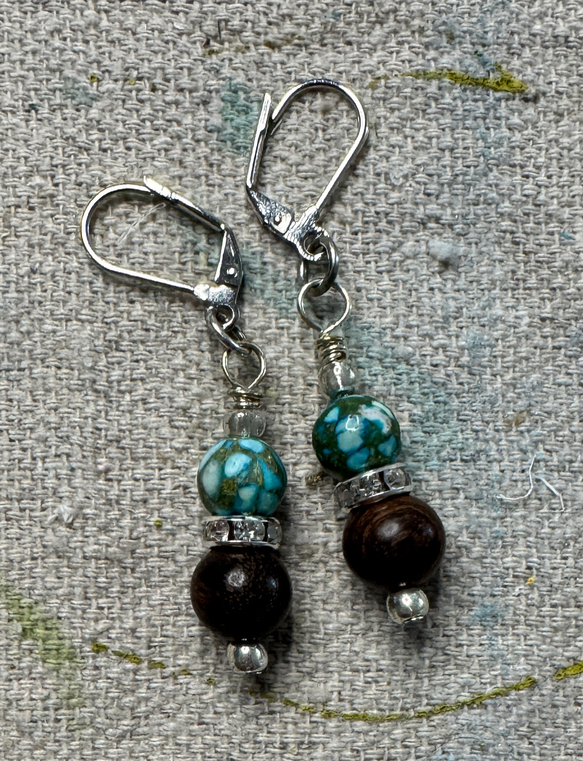 A pair of handmade dangle earrings with turquoise and dark brown beads, displayed on a canvas drop cloth.
