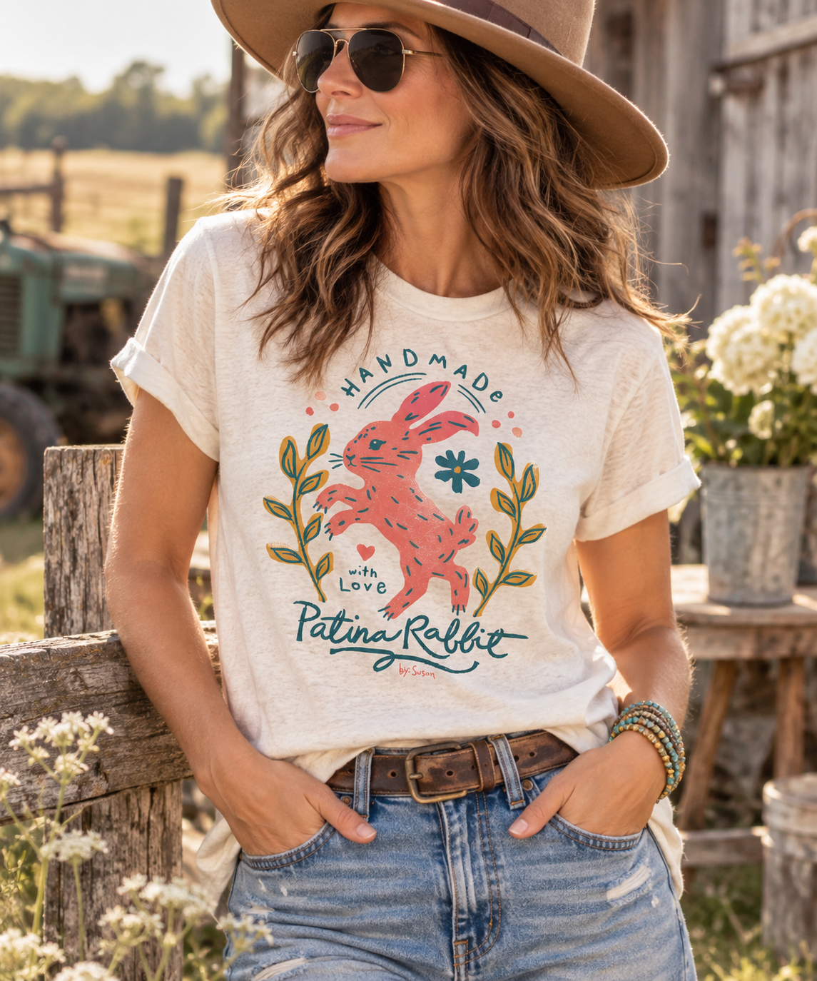 A woman wearing a cream-colored graphic t-shirt with a pink rabbit design and the words 'Handmade with Love Patina Rabbit' stands outdoors in a rustic setting.