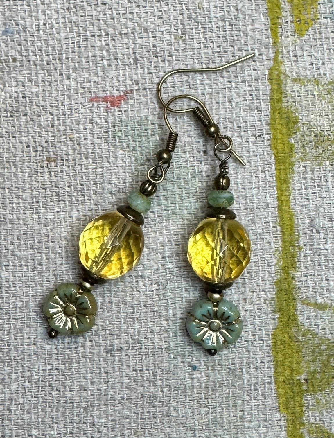 A pair of handmade dangle earrings with faceted lite topaz Czech glass beads, small amazonite beads, Czech Flower with bronze wash, and antiqued bronze-tone metal hooks, displayed on a textured linen fabric background.