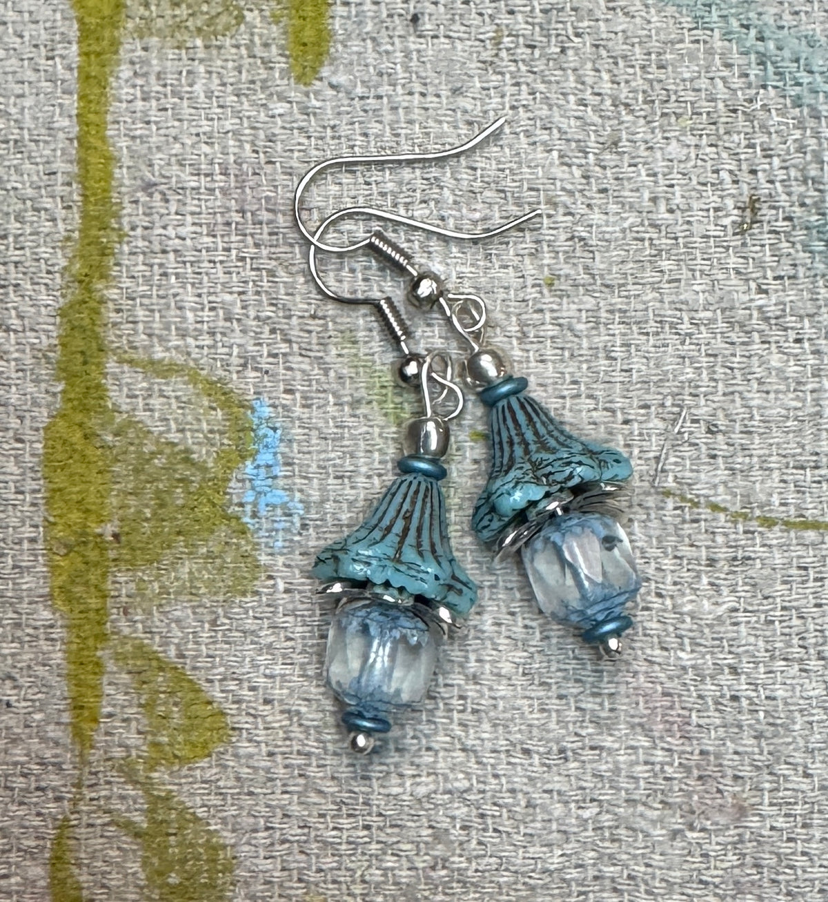 A pair of handmade dangle earrings featuring A pair of bohemian-style dangle earrings  feature weathered Czech, aqua, blue bell shaped beads paired with soft aqua and clear cathedral cut glass beads. Antique silver accents, displayed on a textured beige linen fabric.