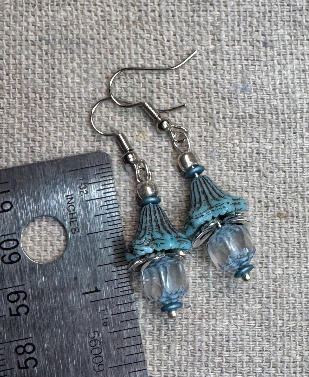 A pair of handmade dangle earrings A pair of bohemian-style dangle earrings  feature weathered Czech, aqua, blue bell shaped beads paired with soft aqua and clear cathedral cut glass beads. Antique silver accents, shown next to a metal ruler on a textured beige fabric background. Ruler show earrings will hang about 1" not including the earwires.