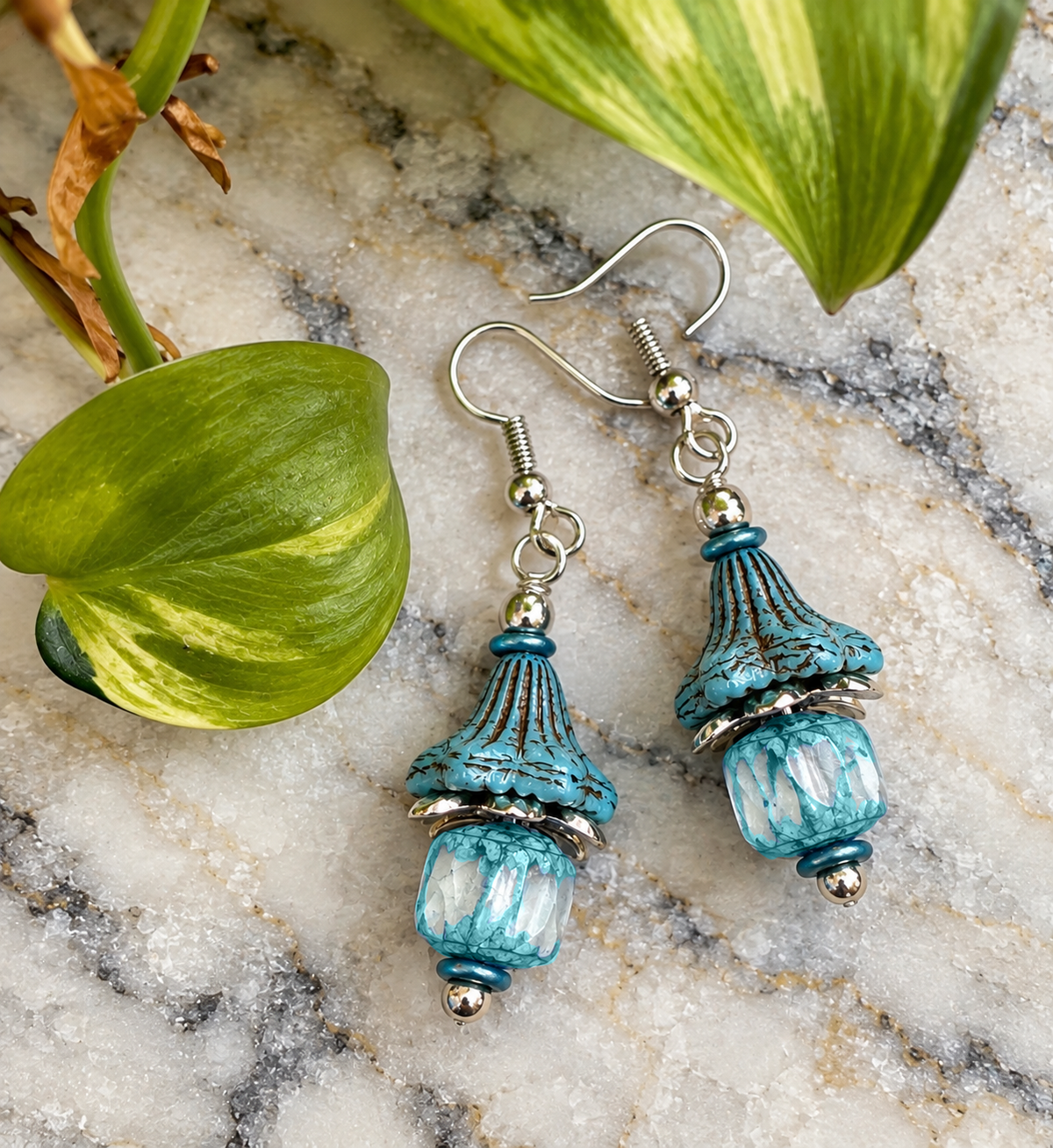 A pair of bohemian-style dangle earrings  feature weathered Czech, aqua, blue bell shaped beads paired with soft aqua and clear cathedral cut glass beads. Antique silver accents, displayed on a white marble surface with green leaves.