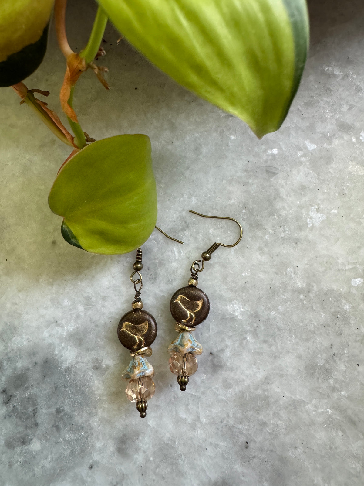 A pair of handmade dangle earrings with dark brown beads, gold-toned metal details, and light gold crystal drops, displayed on a light gray stone surface next to green plant leaves.