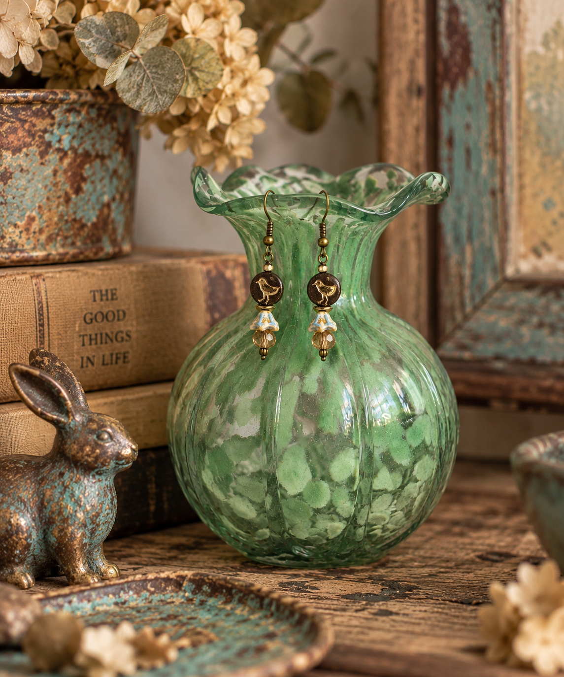 A green marbled glass vase with a scalloped rim sits on a rustic wooden surface, styled with vintage books, a small bronze rabbit figurine, dried flowers, and matching earrings hanging from its rim.