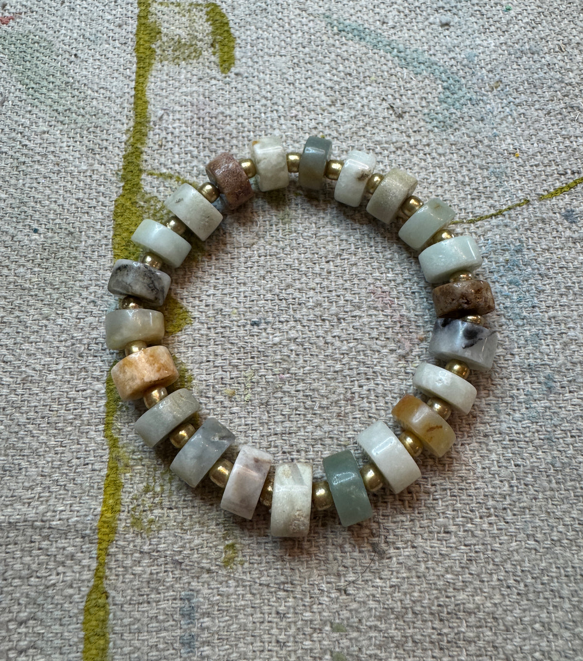 Coastal Stone stretch bracelet featuring 6mm Aqua Amazonite heishi beads in soft aqua, cream, gray and warm natural tones, accented with small gold seed beads sitting on canvas cloth.