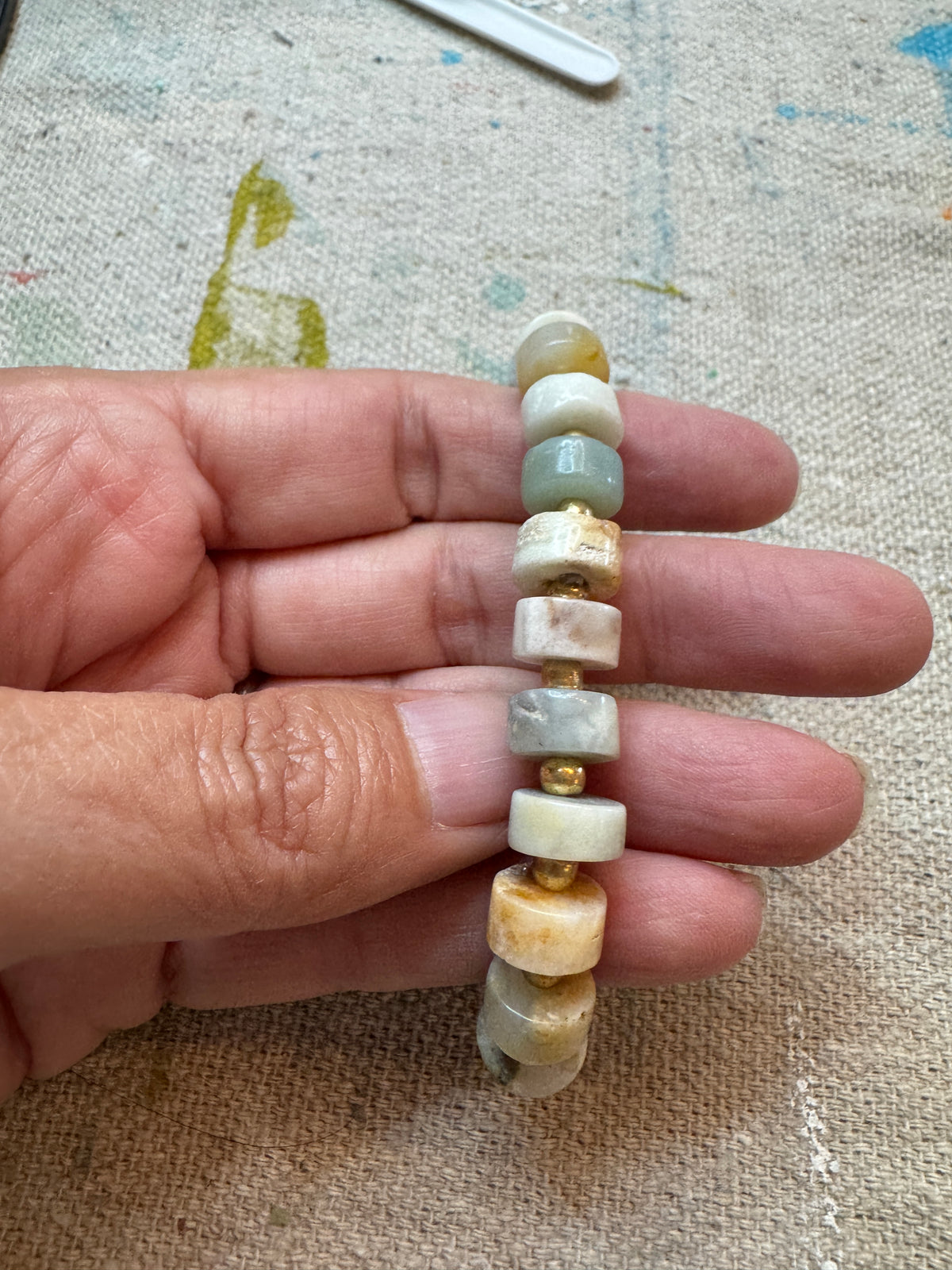 Coastal Stone stretch bracelet featuring 6mm Aqua Amazonite heishi beads in soft aqua, cream, gray and warm natural tones, accented with small gold seed beads. held up in hand .