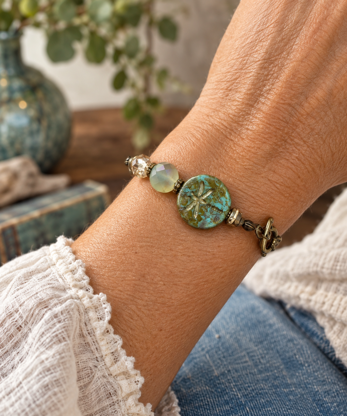 A close-up of a person's wrist wearing a handmade beaded bracelet with a prominent round turquoise dragonfly charm, a light green bead, and bronze-toned metal details.