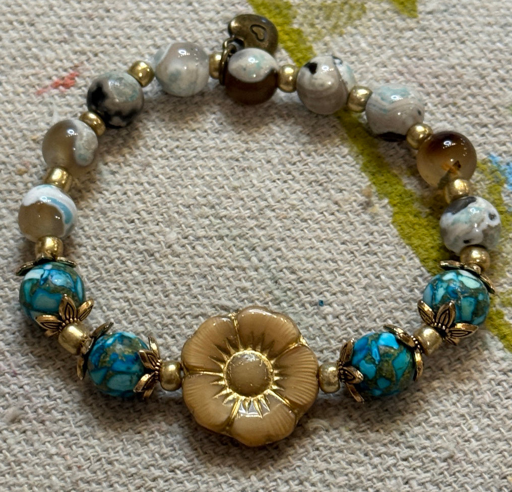 A beaded stretch bracelet featuring a central gold-tone Czech flower charm, turquoise-blue marbled beads, white and brown agate-like beads, and small gold spacer beads, displayed on a textured beige fabric background.