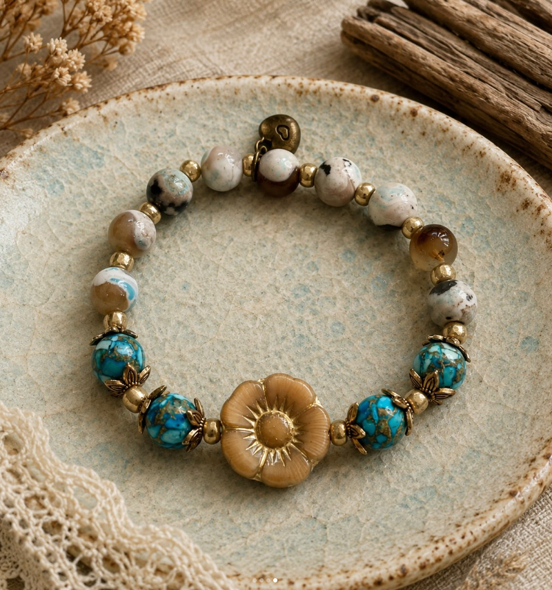 A beaded stretch bracelet with a central gold-tone Czech flower charm, turquoise-blue marbled beads, and white-and-gray stone beads, displayed on a textured ceramic plate.