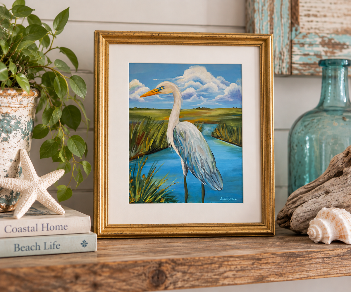 A framed art print depicting a white heron standing in a blue marsh, styled on a wooden shelf with a plant, starfish, books, driftwood, and a glass bottle.