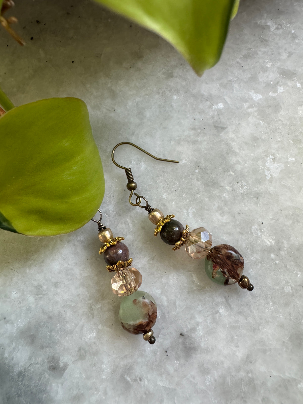 A pair of handmade dangle earrings with bronze-toned metal hooks and beads, featuring faceted clear beads, a dark brown bead, and a larger marbled brown and green bead, displayed on a textured white surface with green leaves.