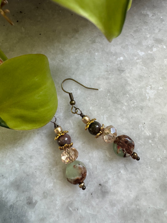 A pair of handmade dangle earrings with bronze-toned metal hooks and beads, featuring faceted clear beads, a dark brown bead, and a larger marbled brown and green bead, displayed on a textured white surface with green leaves.
