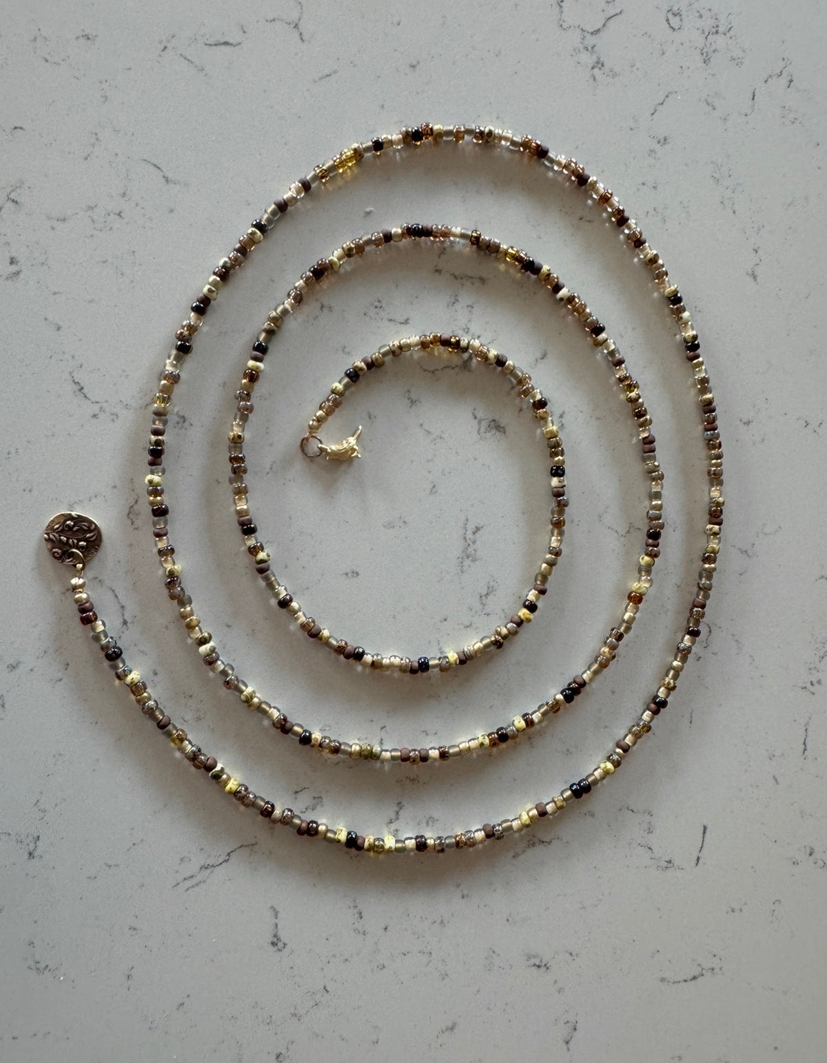 A beaded chain necklace with small metallic beads in gold, silver, and dark bronze tones, arranged in a spiral on a light gray marble-textured surface.