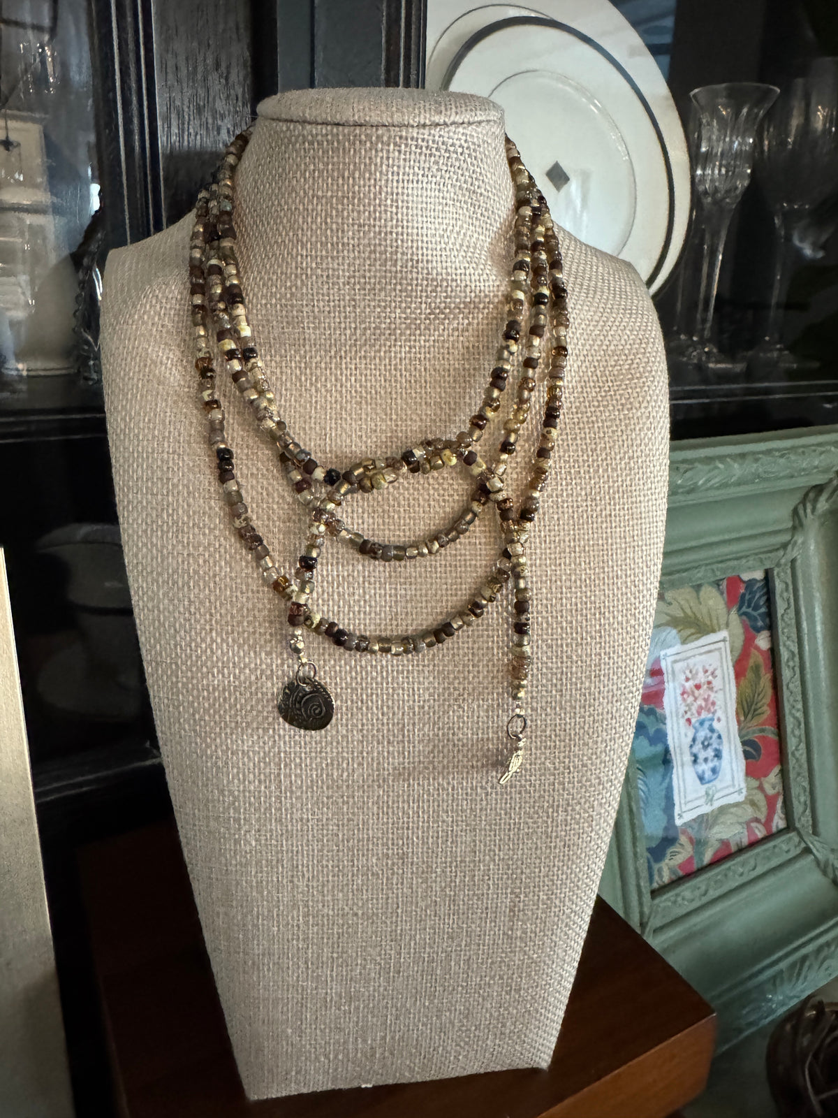 Earthbound 48-inch lariat necklace with earth-tone Picasso seed beads, a gold bird and an antique brass Jardin charm on a Necklace display.