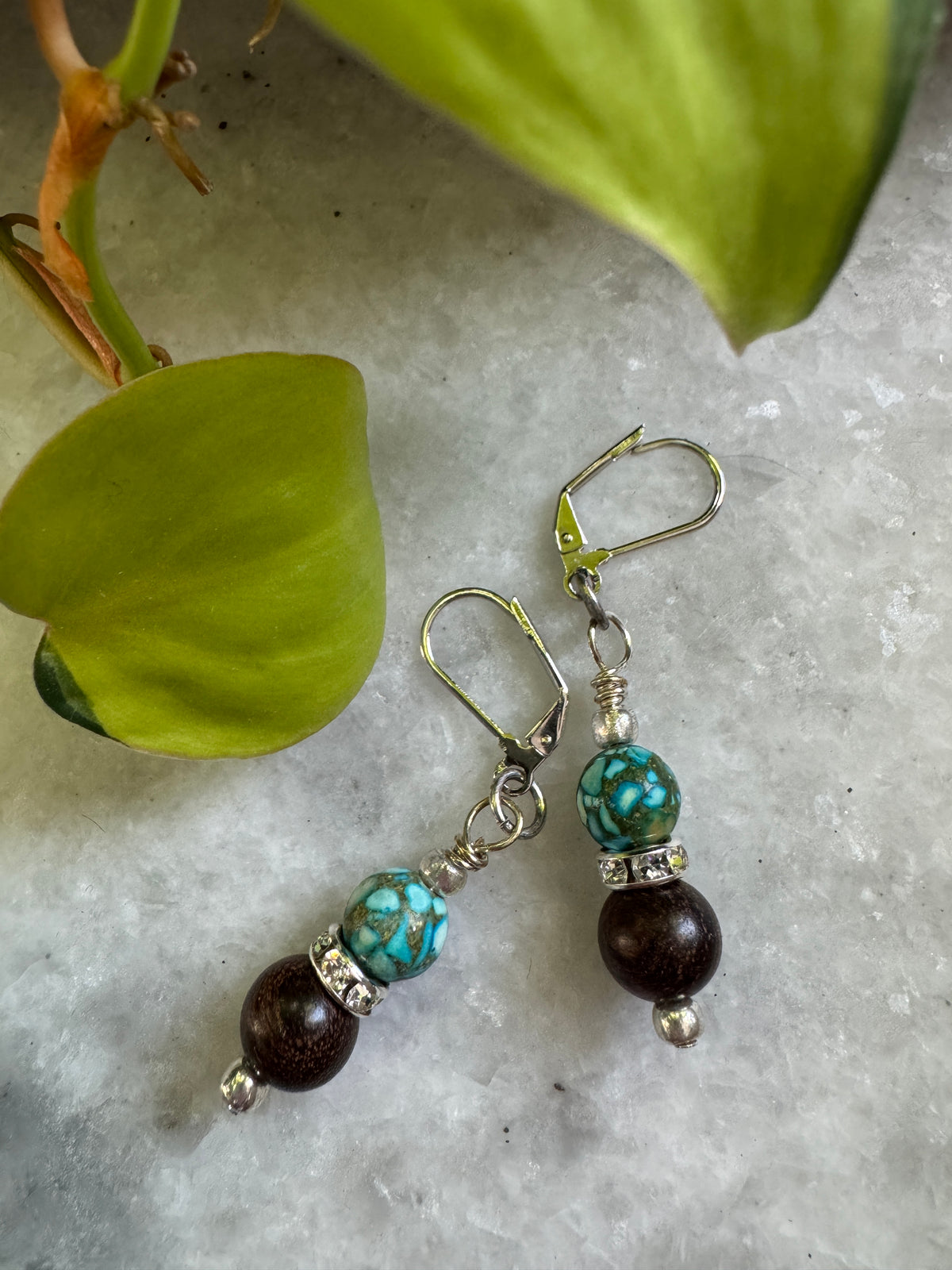 A pair of handmade dangle earrings with turquoise and dark brown beads, displayed on a light gray stone surface next to green leaves.