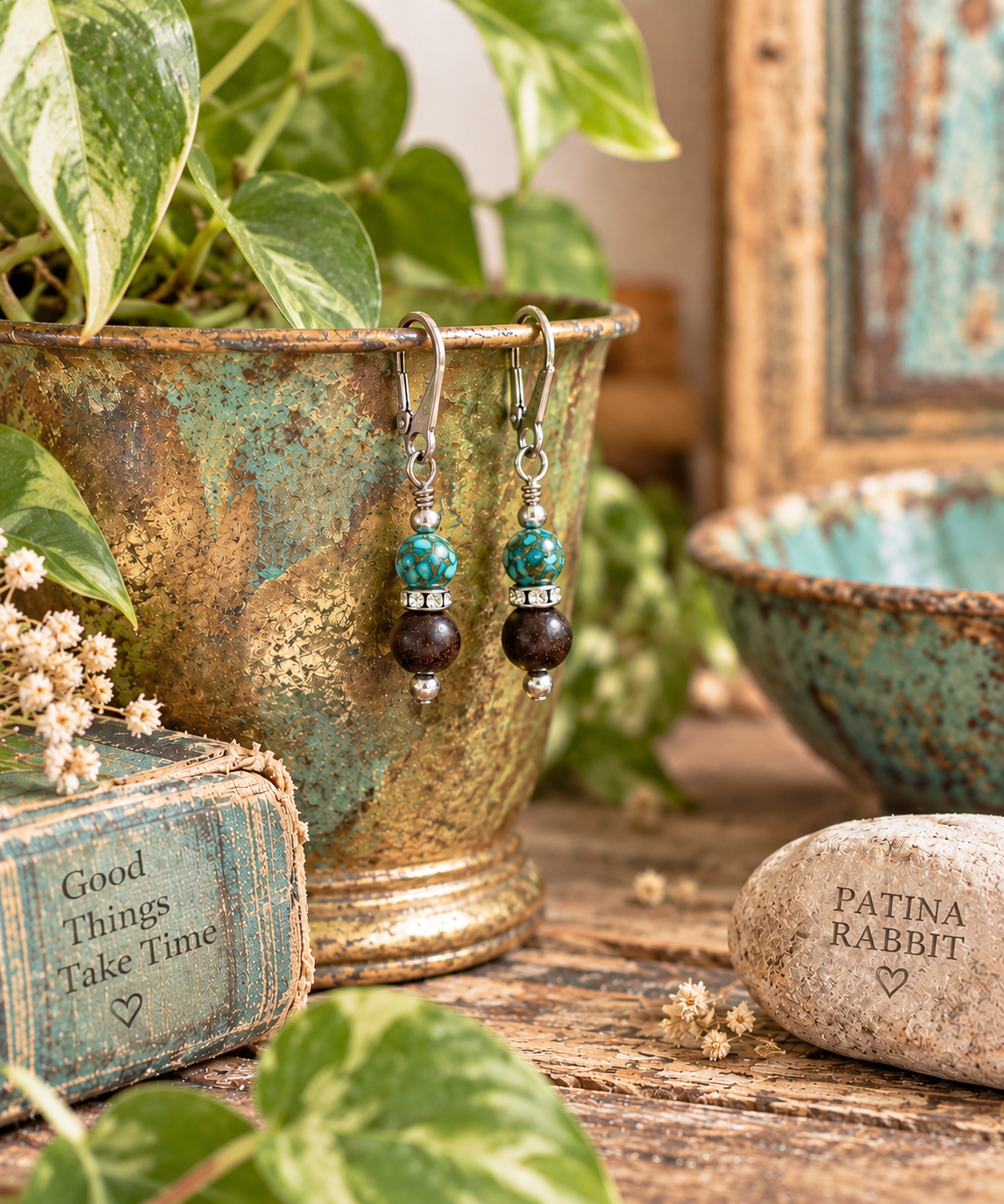 A pair of handmade beaded drop earrings hanging from a rustic bronze pot on a wooden table, styled with green plants, a turquoise bowl, a small book, and a stone engraved with 'PATINA RABBIT'.