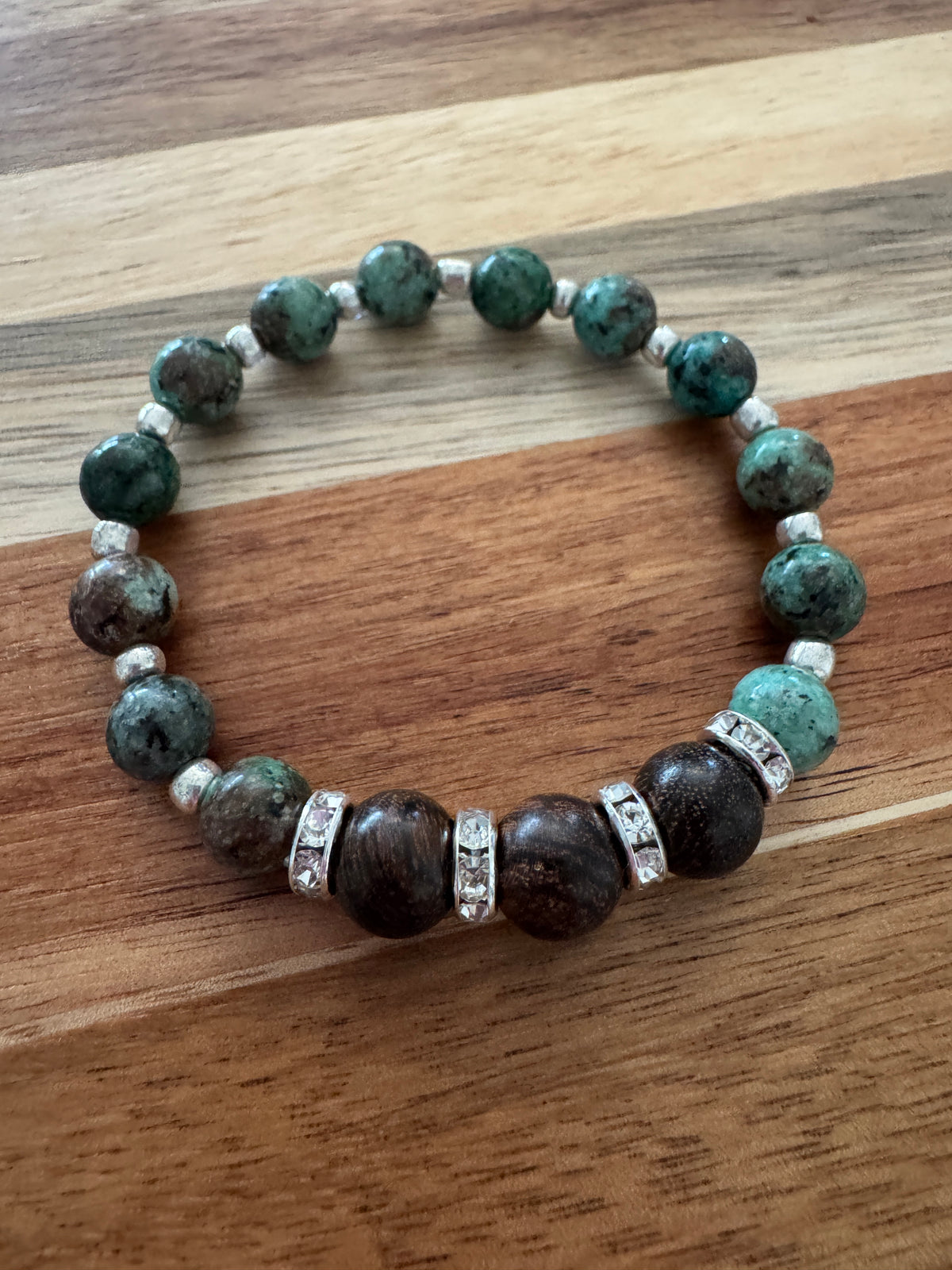 A beaded stretch bracelet featuring dark brown beads and green turquoise marbled beads, silver spacer beads, and silver-toned metal spacers with clear stones, displayed on a wooden surface.