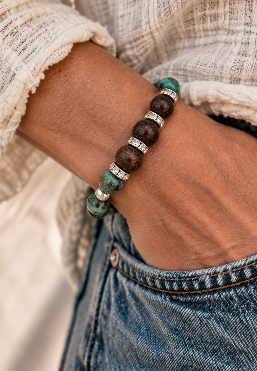 A beaded stretch bracelet featuring dark brown beads and green turquoise marbled beads, silver spacer beads, and silver-toned metal spacers with clear stones, shown on a wrist with hand in blue jeans.