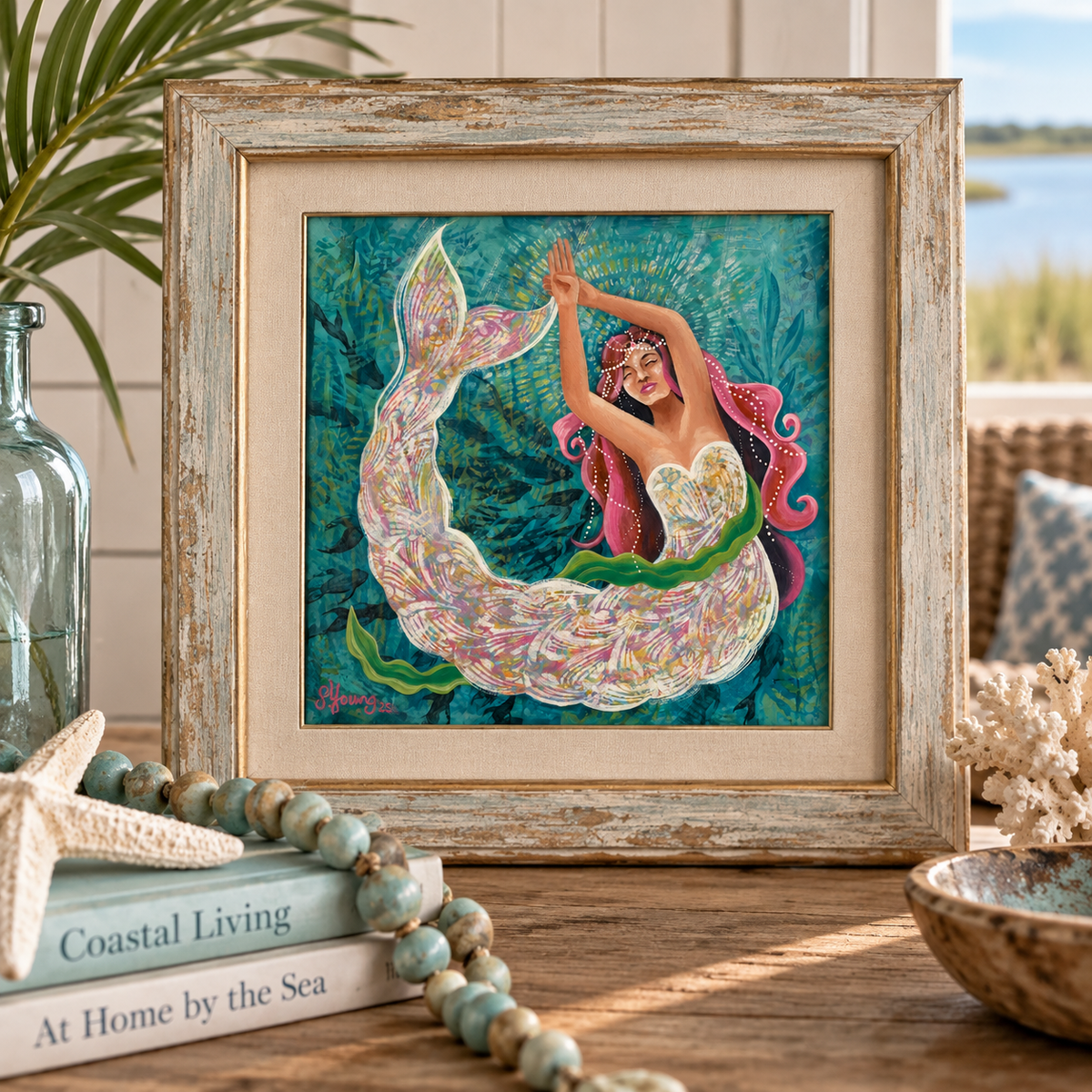 A framed art print featuring a colorful mermaid illustration, styled on a wooden table with coastal decor including a glass bottle, starfish, beads, and a bowl.