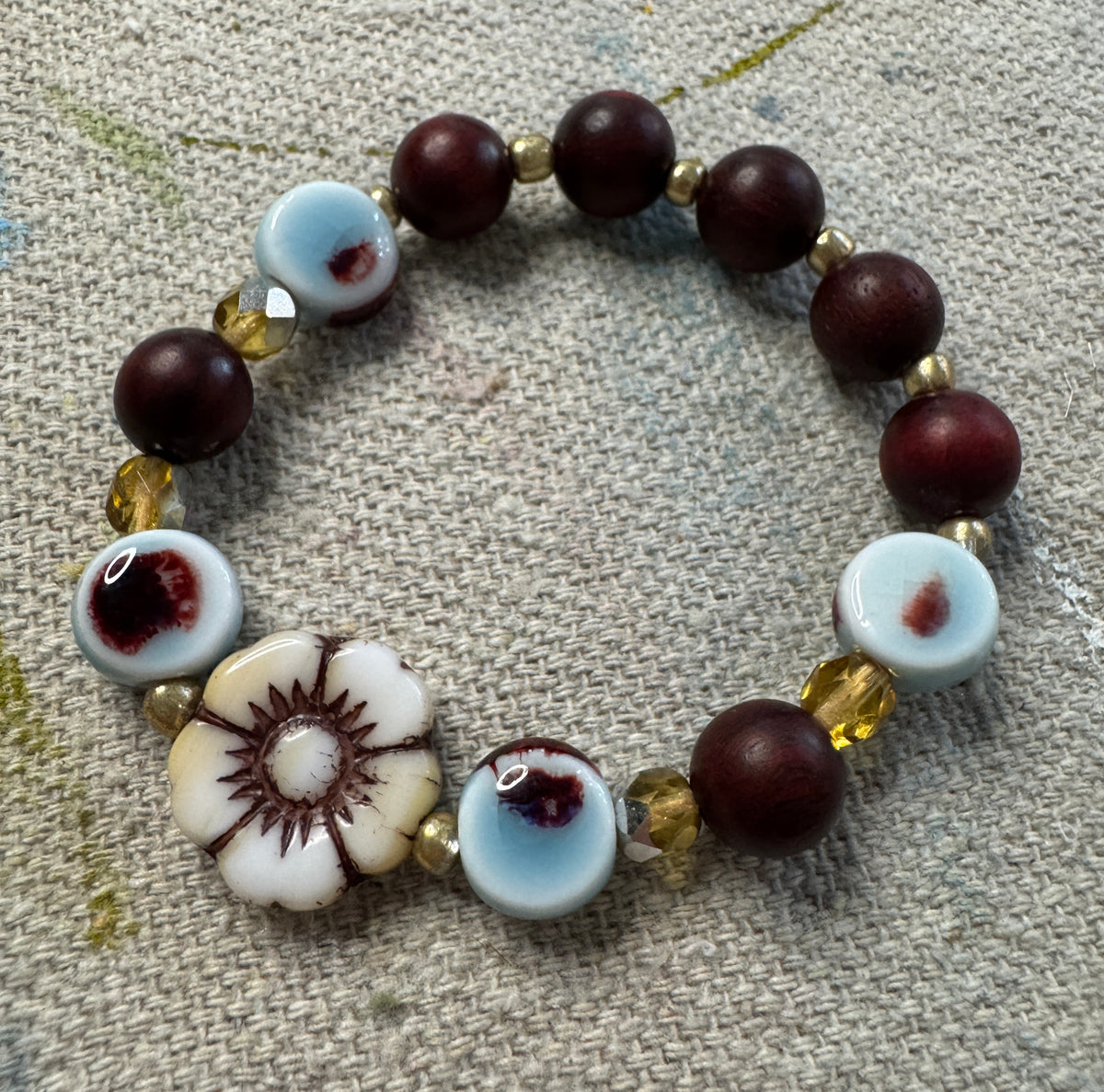 A handmade beaded bracelet featuring dark reddish-brown wooden beads, light blue and white ceramic beads with dark red spots, a white Czech flower bead with brown details, and small faceted yellow beads, displayed on a textured gray fabric background.