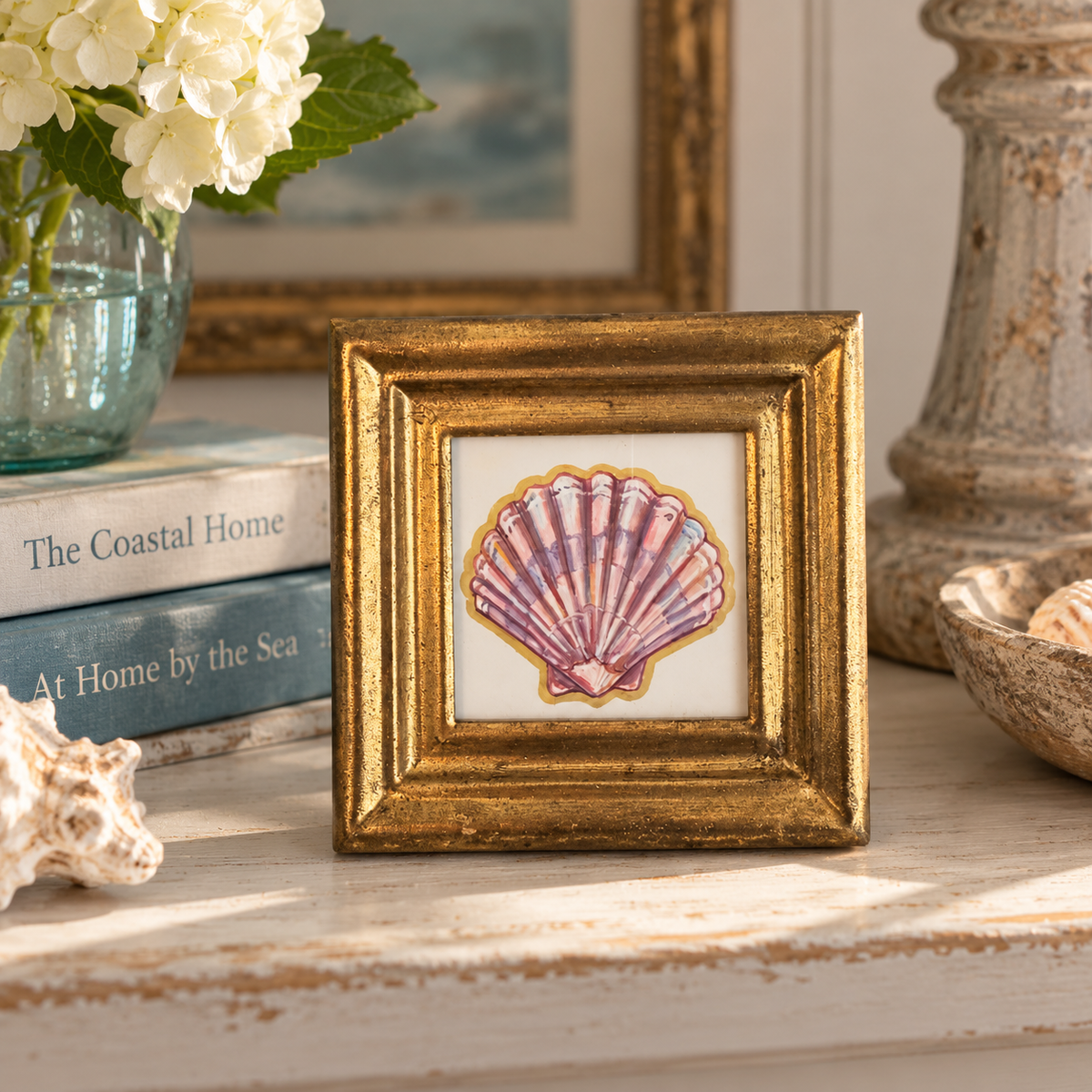 A small framed art print featuring a pink and purple seashell on a white background, set in a distressed gold frame. The frame is styled on a rustic white wooden table with a blue glass vase of white flowers, a stack of books, and a seashell bowl nearby.