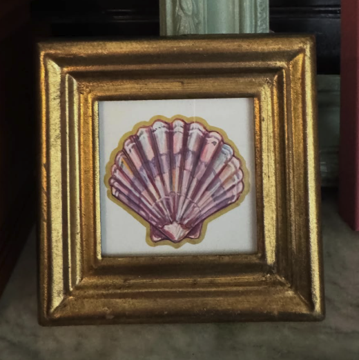 A square gold picture frame containing a painted pink, purple and gold seashell illustration on a white background, photographed indoors against a dark and light background.