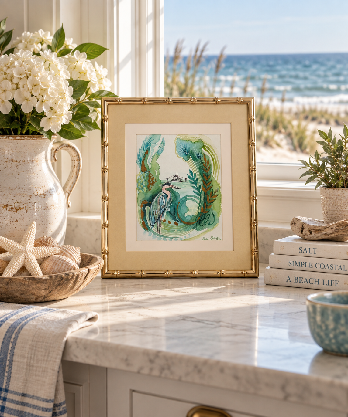 A framed watercolor painting featuring a whimsical illustration of a heron and shrimp boat in the distance in green, blue, and white tones, styled on a marble kitchen counter with a beach view in the background.