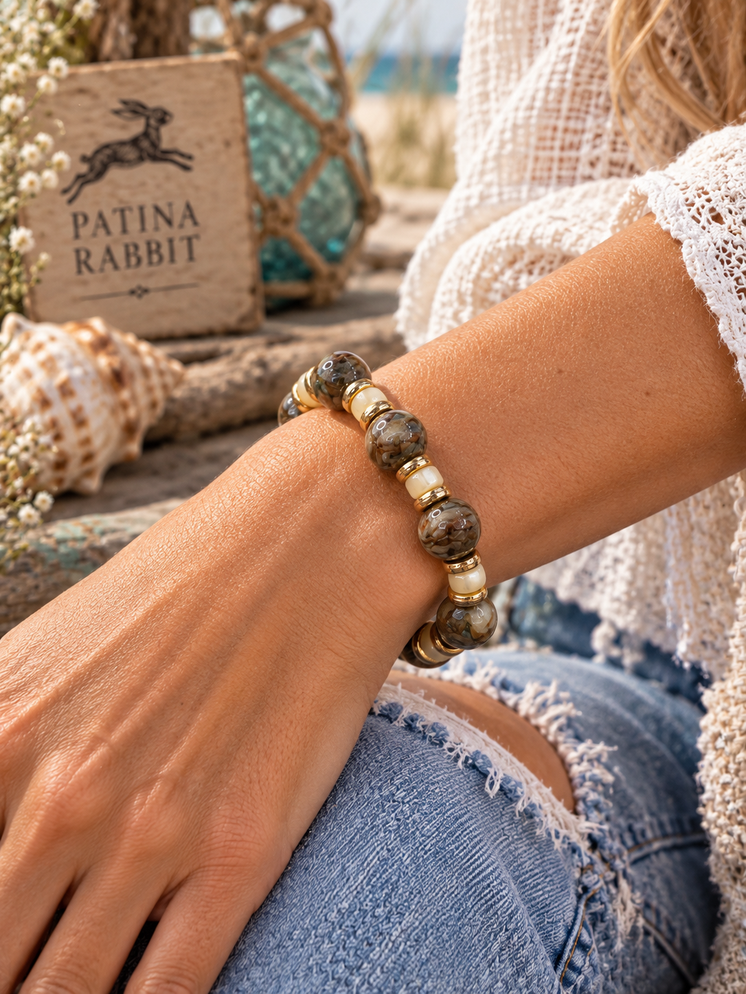 A close-up lifestyle shot of a woman's wrist wearing a beaded bracelet made of brown and beige Lampwork stone beads with small pearl heishi beads and gold-toned spacer beads. She is wearing a white lace top and ripped blue jeans, with a 'PATINA RABBIT' wooden sign and seashells in the background.